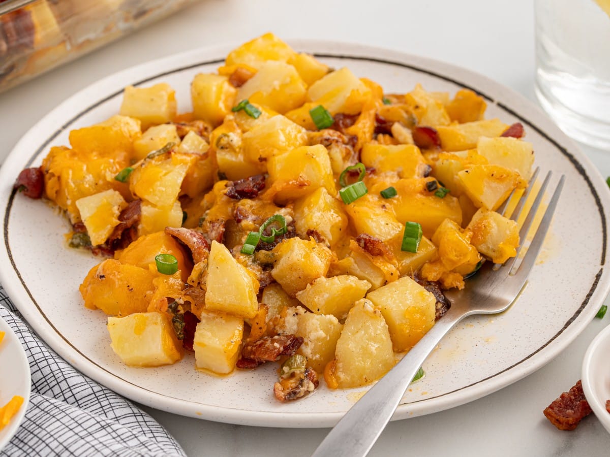 Serving of Mississippi Mud Potatoes on a speckled plate with cubed potatoes, melted cheddar cheese, bacon pieces, and green onions, served with a fork.
