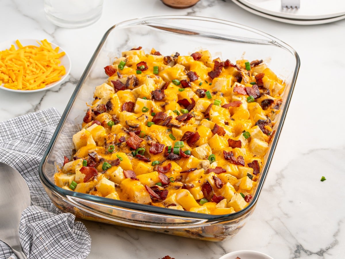 Baked Mississippi Mud Potatoes in a clear glass casserole dish with cubed Yukon gold potatoes, melted cheddar cheese, bacon, and green onions on a marble countertop.