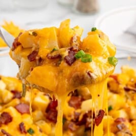 Close-up of Mississippi Mud Potatoes with melted cheddar cheese, bacon pieces, and green onions being lifted from the baking dish with a serving spoon.