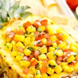 Fresh pineapple salsa in a hollowed pineapple half with juicy diced pineapple, tomatoes, red onion, cilantro, and tortilla chips, with lime and fresh produce in the background.