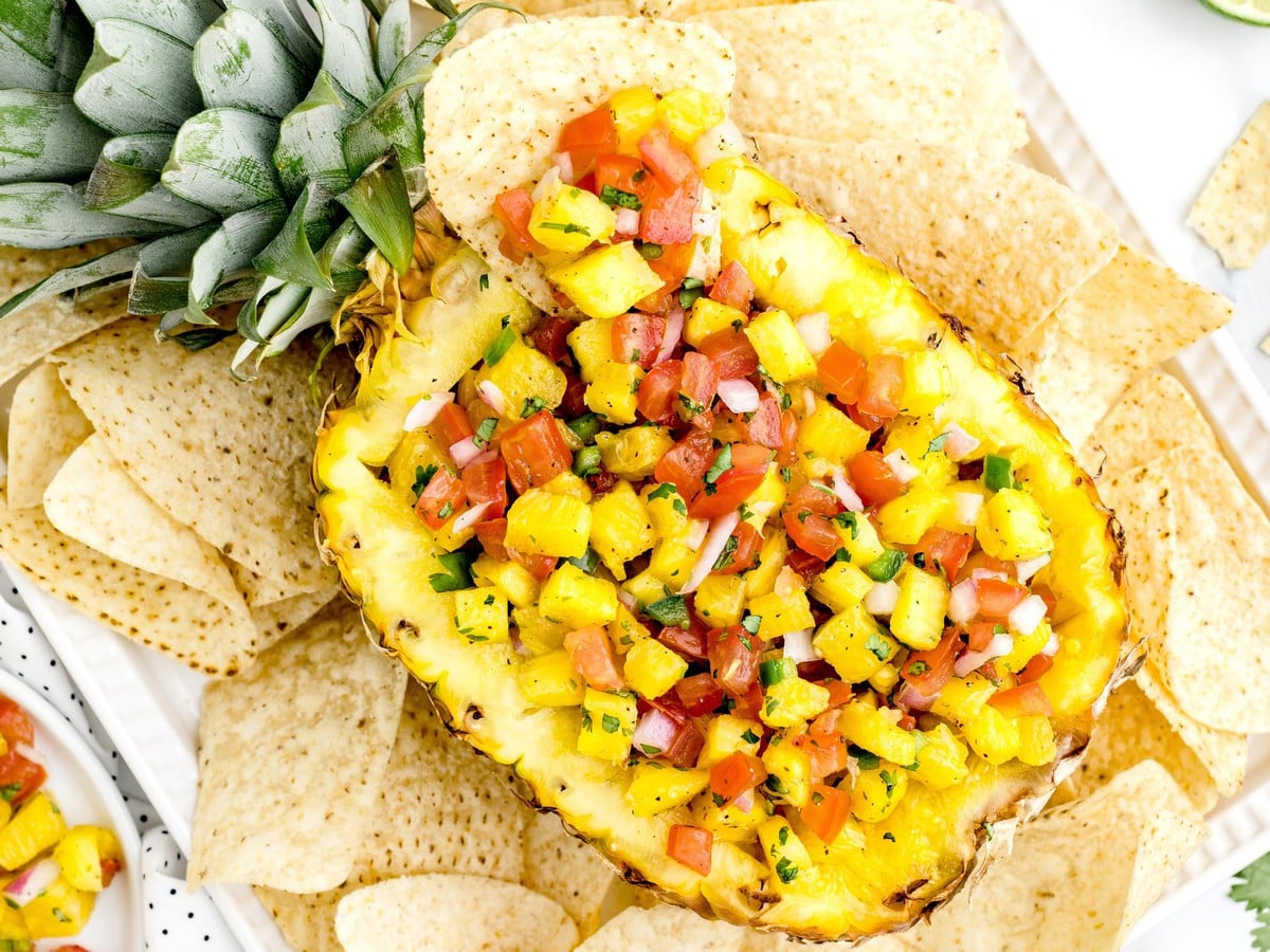 Overhead view of fresh pineapple salsa served in a hollowed pineapple half, filled with diced pineapple, tomatoes, red onion, jalapeño, and cilantro, surrounded by tortilla chips.