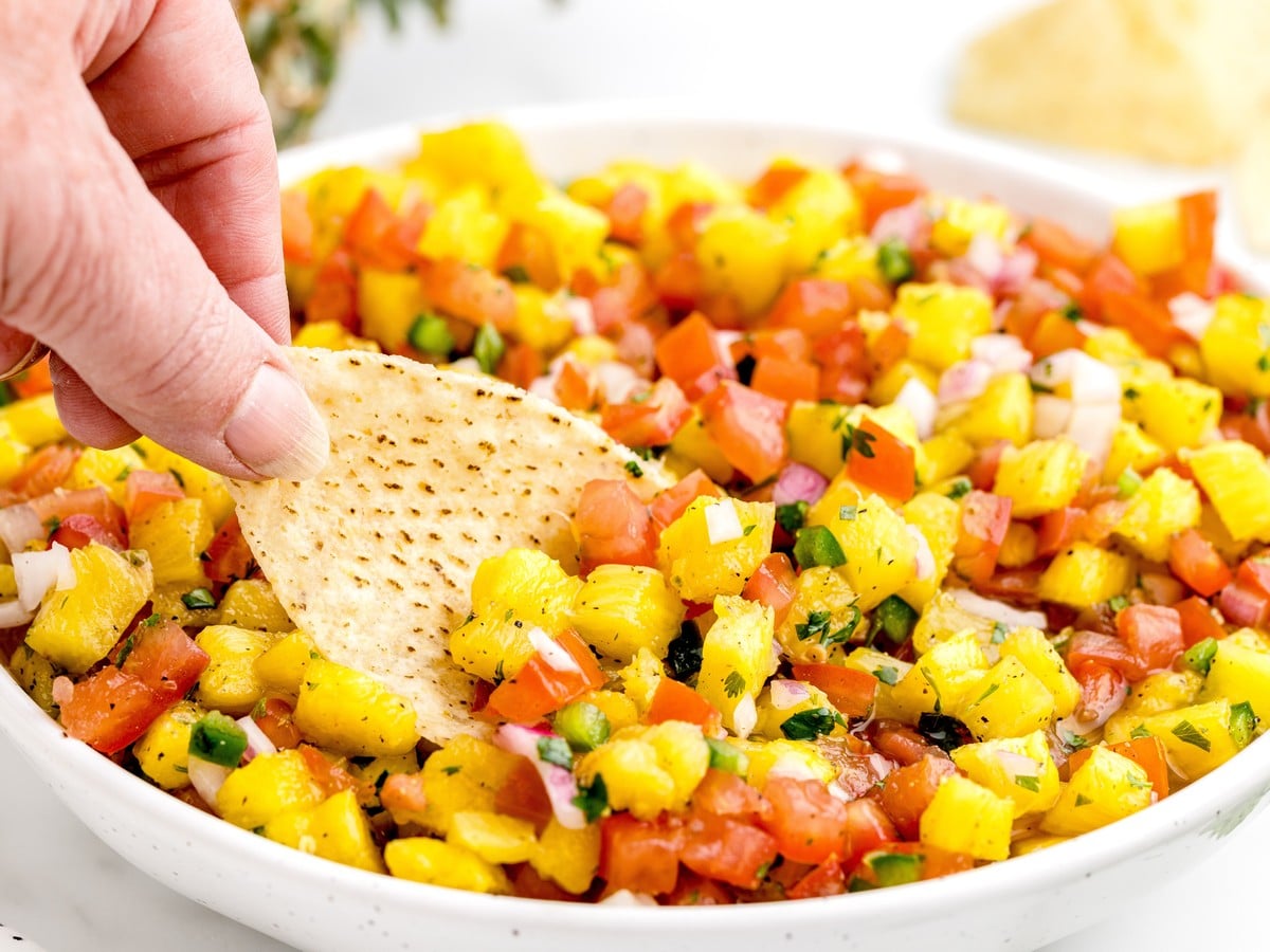 Close-up of fresh homemade pineapple salsa in a white bowl with diced pineapple, tomatoes, red onion, jalapeño, and cilantro, with a tortilla chip being dipped into the salsa.