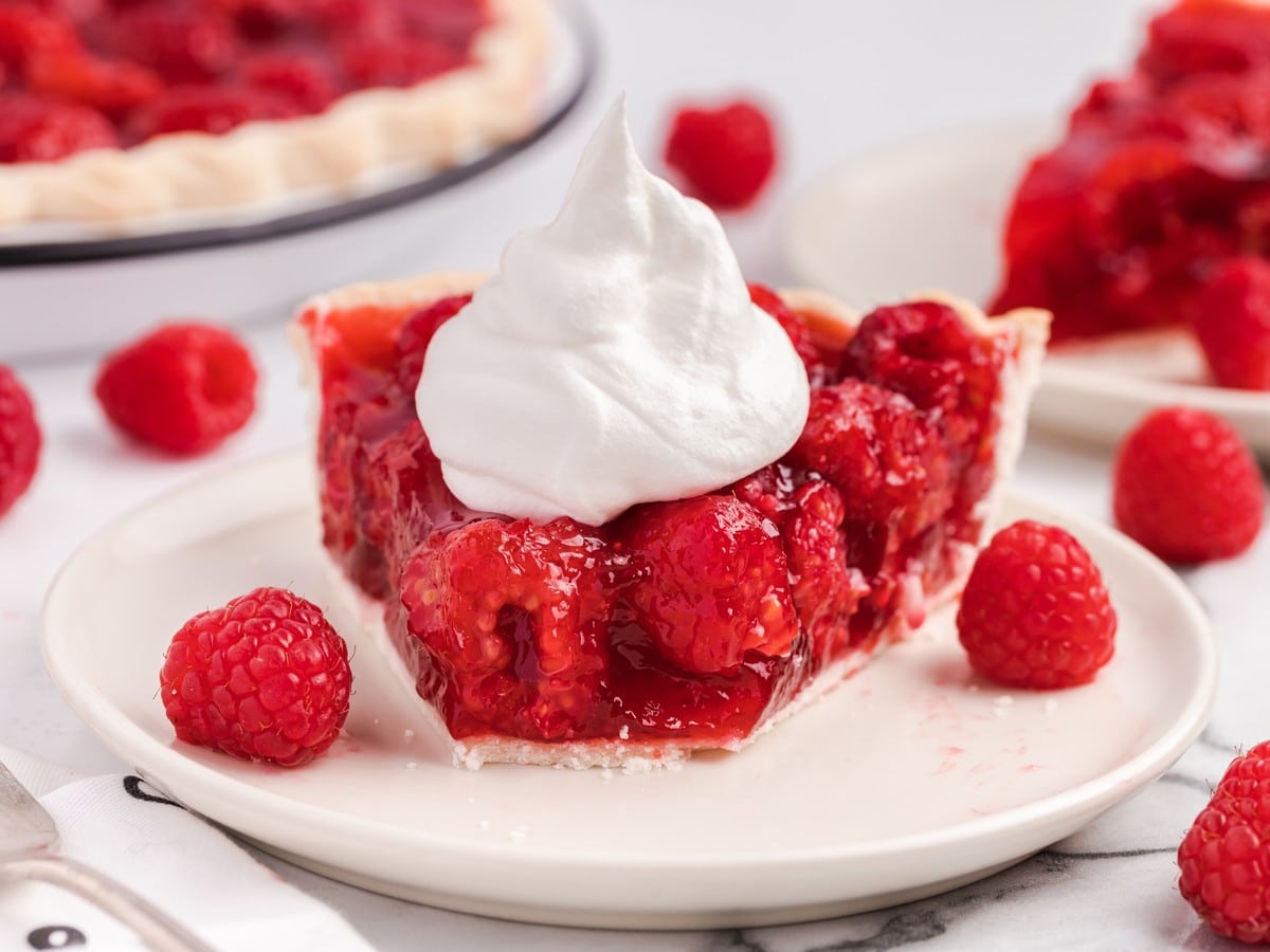 Slice of raspberry pie with glossy red raspberry filling, flaky crust, whipped cream topping, and fresh raspberries on a white plate.