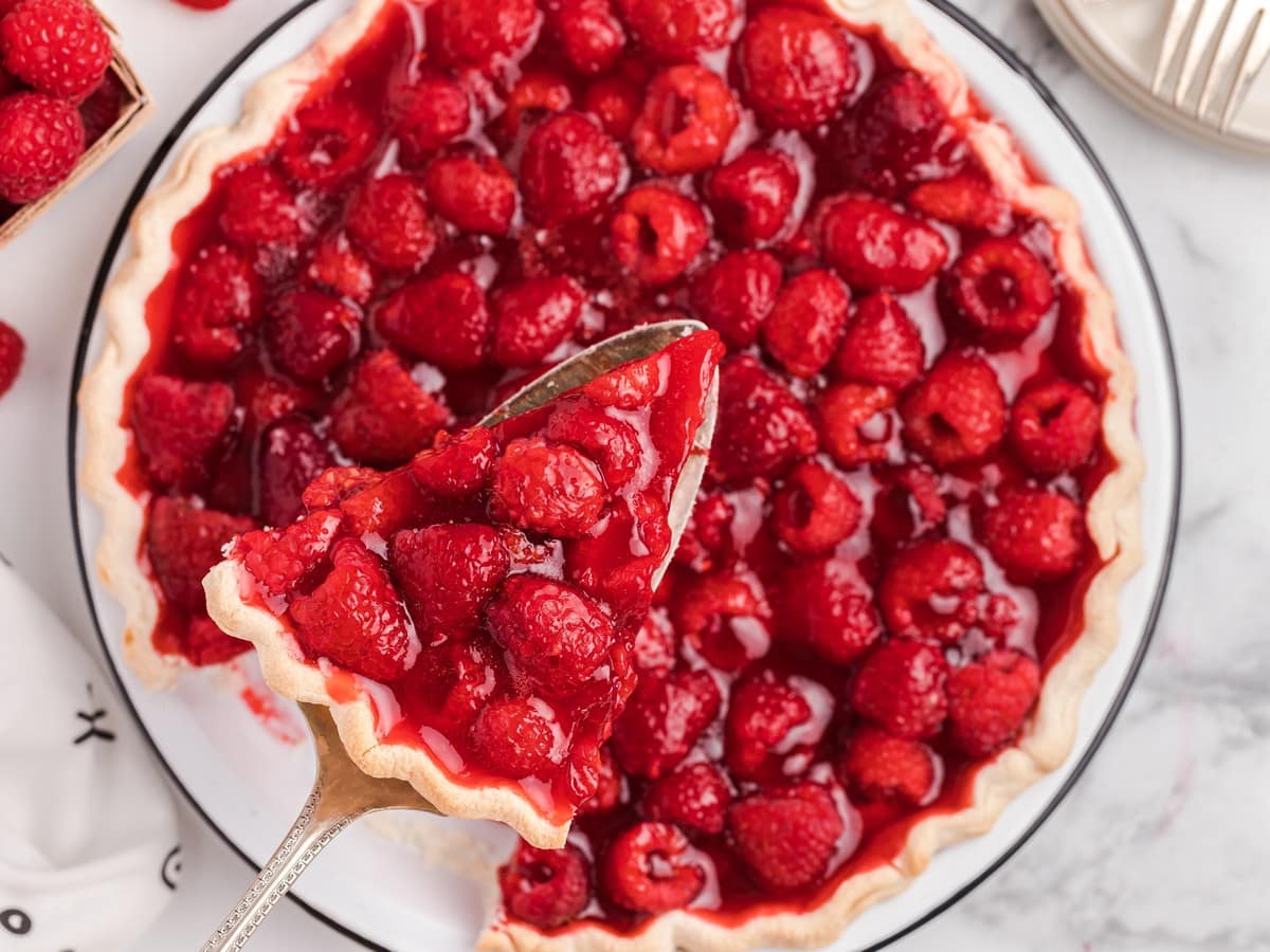 Overhead view of homemade raspberry pie filled with fresh raspberries in a glossy raspberry glaze, with a pie server lifting a generous slice from the flaky crust.