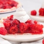 Close-up side view of raspberry pie slice with glossy raspberry filling packed with whole fresh raspberries, flaky crust, whipped cream topping, and fresh raspberries on a white plate.