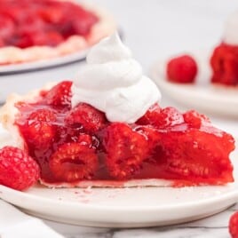 Close-up side view of raspberry pie slice with glossy raspberry filling packed with whole fresh raspberries, flaky crust, whipped cream topping, and fresh raspberries on a white plate.
