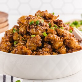 Sesame chicken piled in a white bowl with glossy sauce, sesame seeds, chopped green onions, and chopsticks on the side.