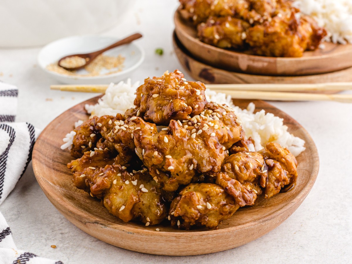 Sesame chicken served over white rice in a wooden bowl with glossy sauce, sesame seeds, and crispy chicken pieces.
