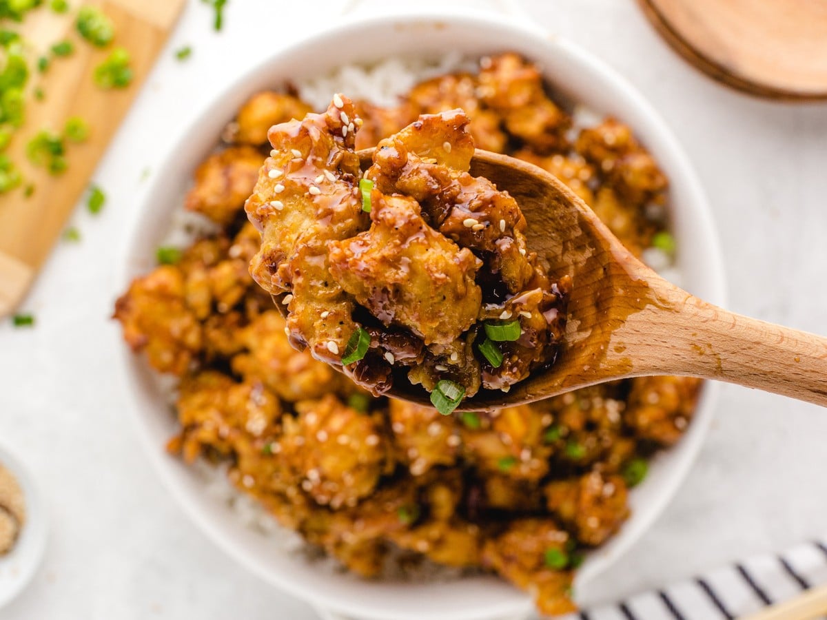 Close-up of crispy sesame chicken coated in glossy sticky sauce with sesame seeds and green onions, served over white rice on a wooden spoon above the bowl.
