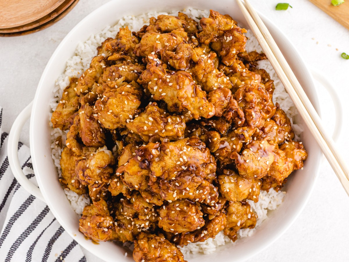 Overhead view of homemade sesame chicken with crispy golden chicken pieces coated in glossy sesame sauce, sprinkled with sesame seeds, and served over fluffy white rice with chopsticks.