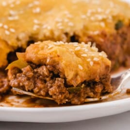 Close-up side view of a slice of sloppy joe bake served on a white plate, featuring layers of flaky golden crust, melted cheddar cheese, and savory sloppy joe ground beef filling.