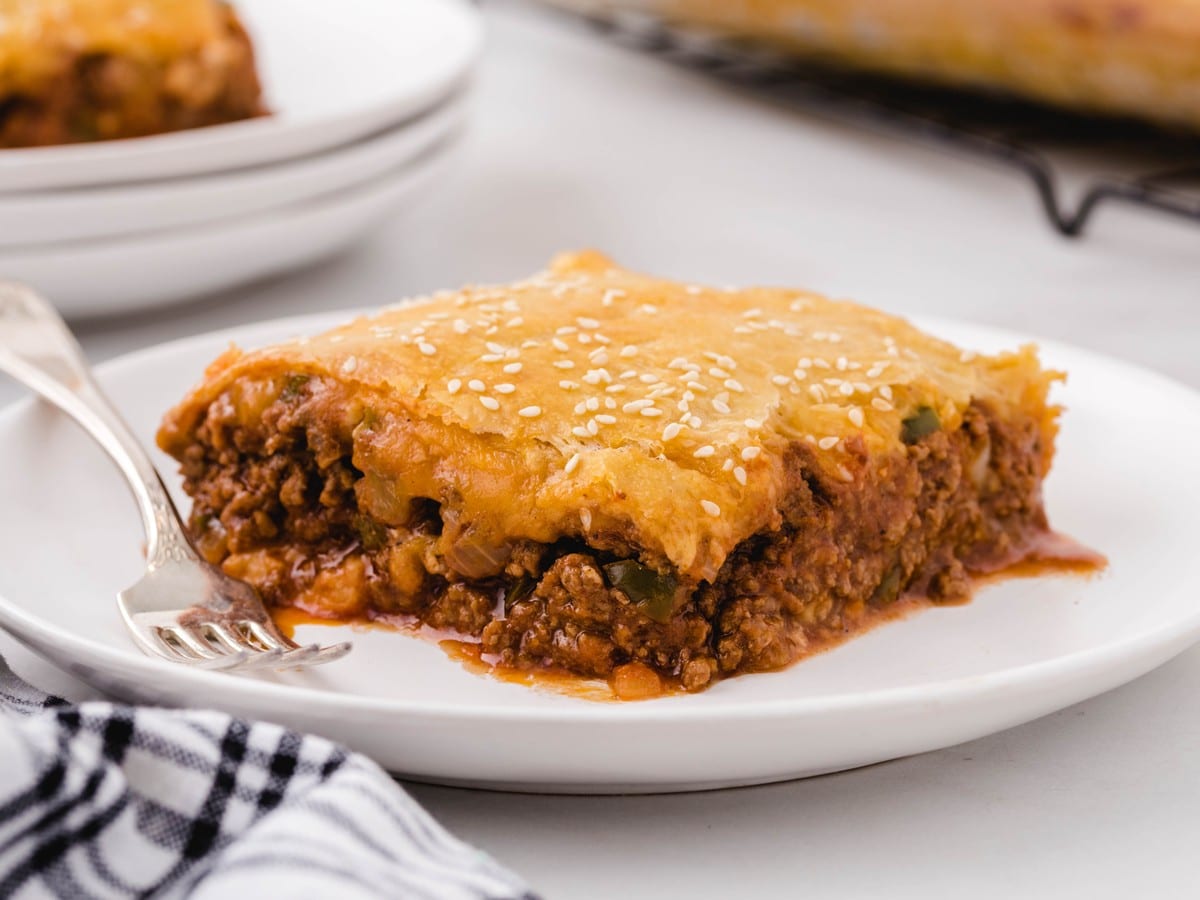 Close-up side view of a slice of homemade sloppy joe bake on a white plate, featuring layers of flaky golden crescent roll crust, melted cheddar cheese, seasoned ground beef, onions, green peppers, and savory sloppy joe sauce.
