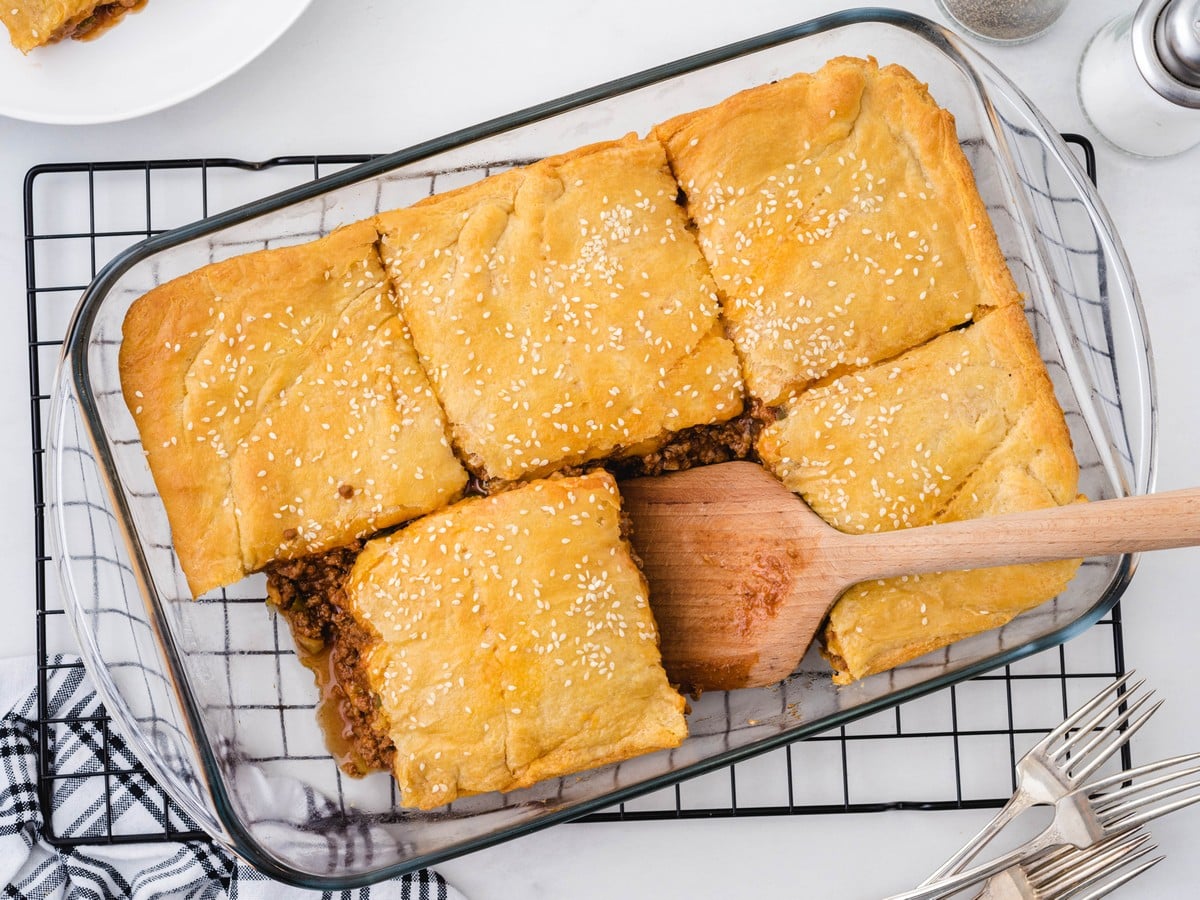 Overhead view of homemade sloppy joe bake in a glass baking dish, topped with golden flaky biscuit crust and sesame seeds, with one square lifted out to show the cheesy seasoned ground beef filling.