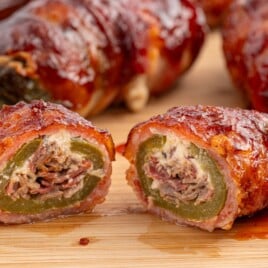 Close-up of sliced Texas Twinkies on a wooden board showing smoked brisket and creamy cheese stuffed inside jalapenos wrapped in bacon and coated with sticky barbecue sauce.
