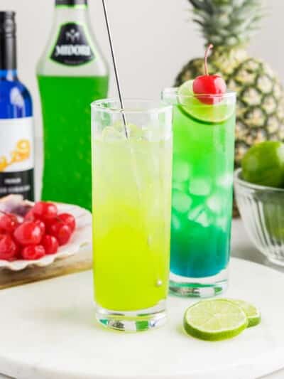 Glass of layered Tipsy Mermaid cocktails with bright green and blue colors, served over ice and garnished with lime slices and maraschino cherries, with bottles of Midori and blue cura&ccedil;ao in the background.