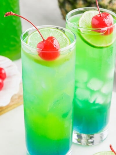 Two layered Tipsy Mermaid cocktails with bright green and blue colors, served over ice and garnished with lime slices and maraschino cherries.