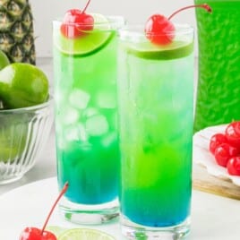 Tipsy Mermaid rum cocktail served in tall glasses with bright layered tropical colors, ice cubes, lime garnish, and maraschino cherries.