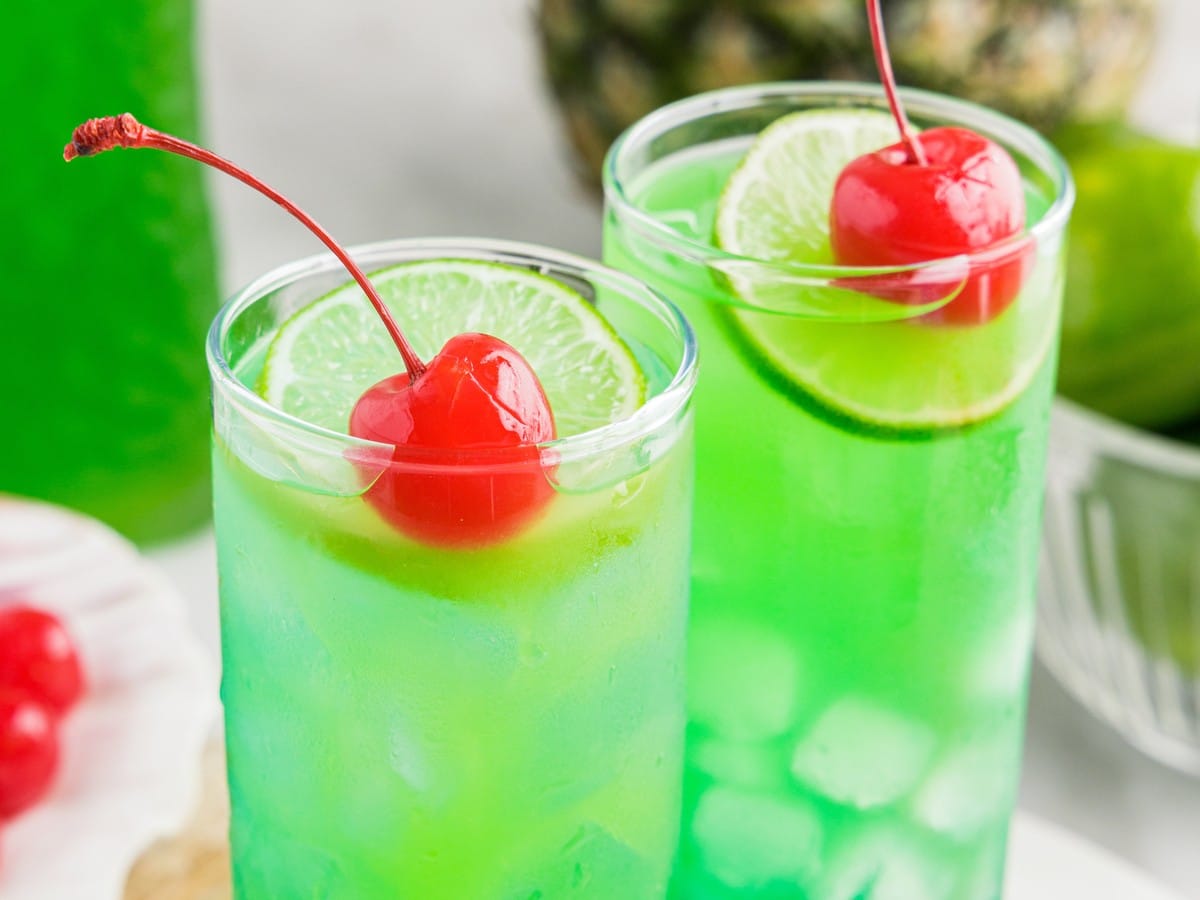 Close-up of Tipsy Mermaid cocktail glasses with ice, lime slices, bright tropical blue and green layers, and maraschino cherry garnish.