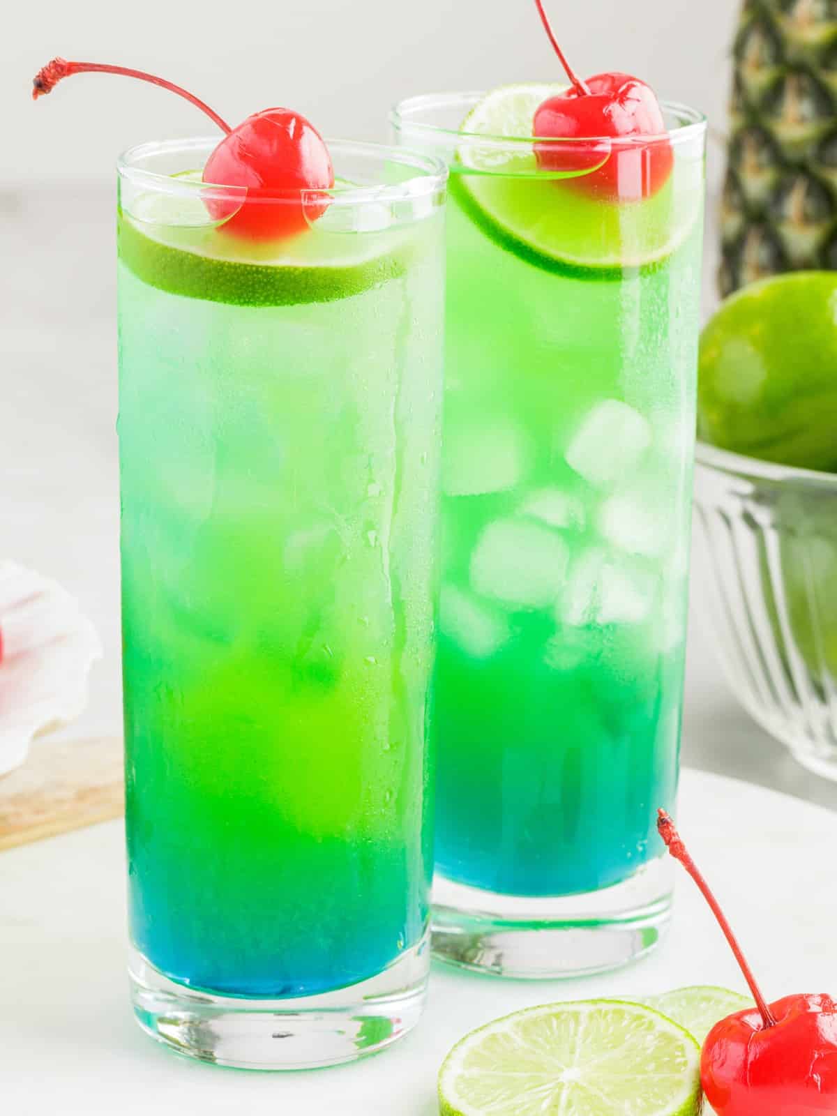 Tall Tipsy Mermaid tropical cocktails with layered blue and green rum drink colors, ice, lime slices, and maraschino cherries on a bright serving setup.