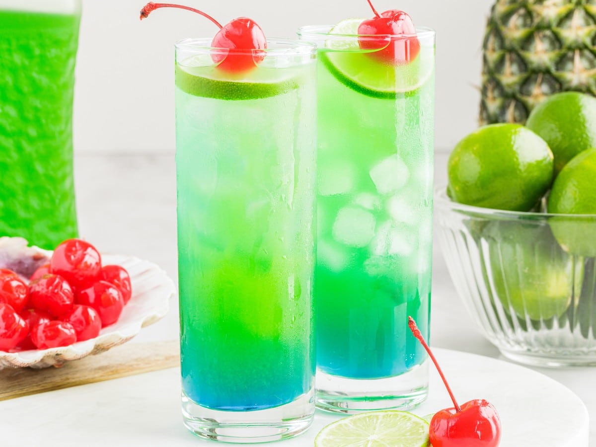 Tipsy Mermaid cocktail in tall glasses with layered blue and green tropical colors, ice, lime slices, and maraschino cherry garnish.