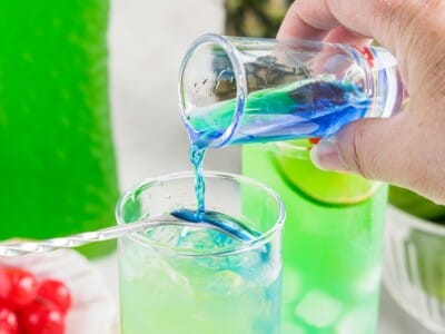 Hand pouring blue cura&ccedil;ao over a spoon into a glass of Tipsy Mermaid cocktail to create the layered blue and green effect.