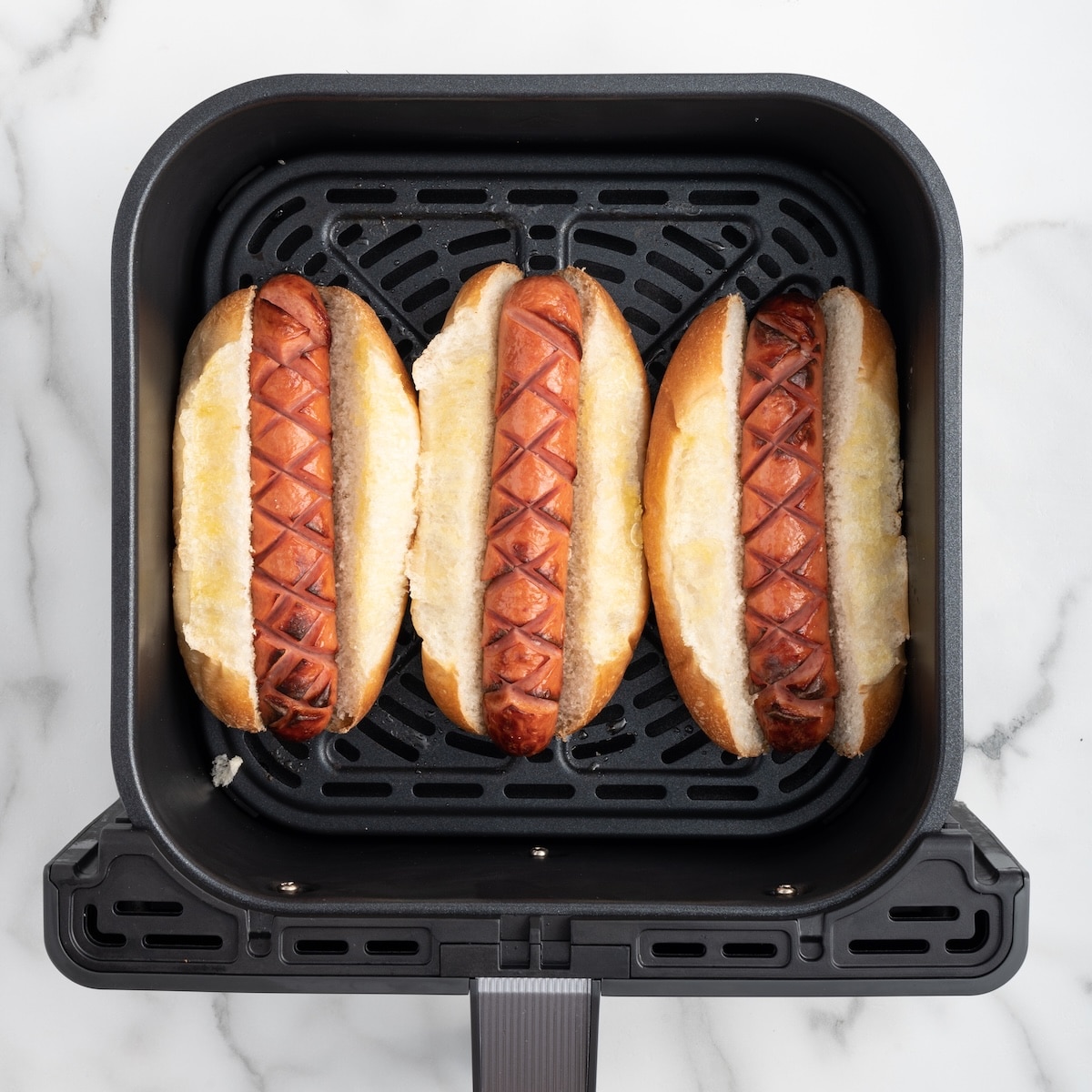 Three air fryer hot dogs in toasted buttered buns in an air fryer basket, ready to serve.