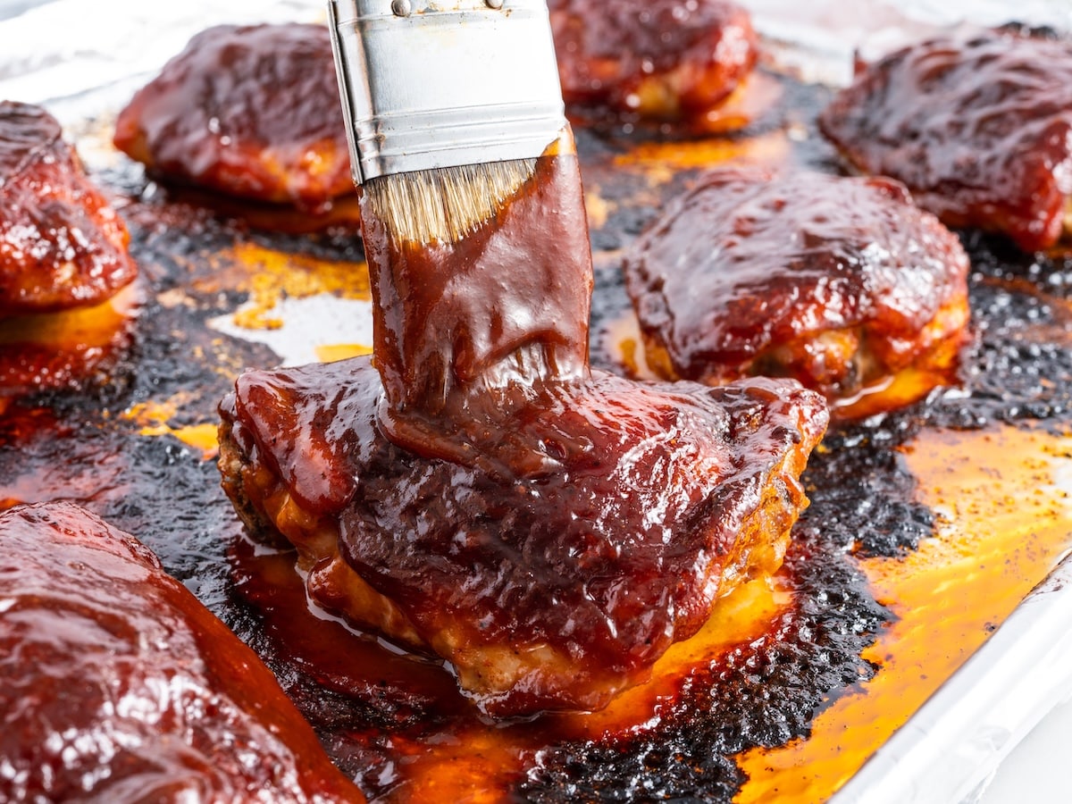 A pastry brush applying a thick layer of BBQ sauce over baked chicken thighs on a foil-lined baking sheet.