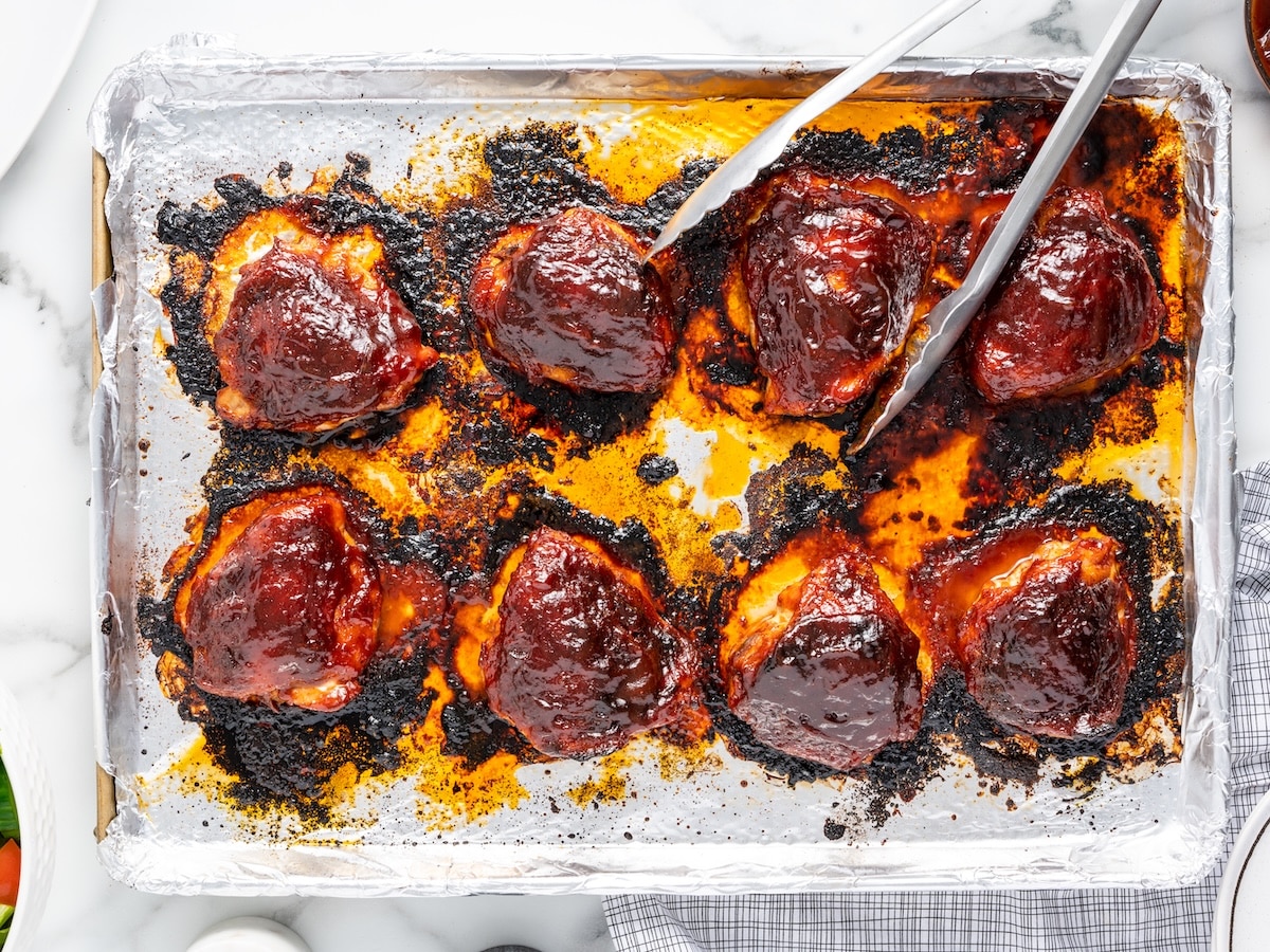 Eight fully baked and broiled BBQ chicken thighs on a foil-lined baking sheet with caramelized glaze.