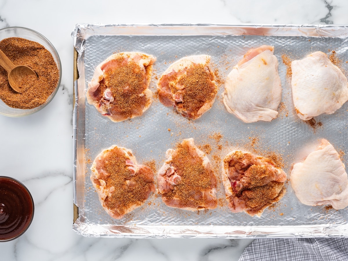 Eight bone-in chicken thighs on a foil-lined baking sheet, half coated in a smoky spice rub, with a bowl of spice rub and a bowl of BBQ sauce on the side.