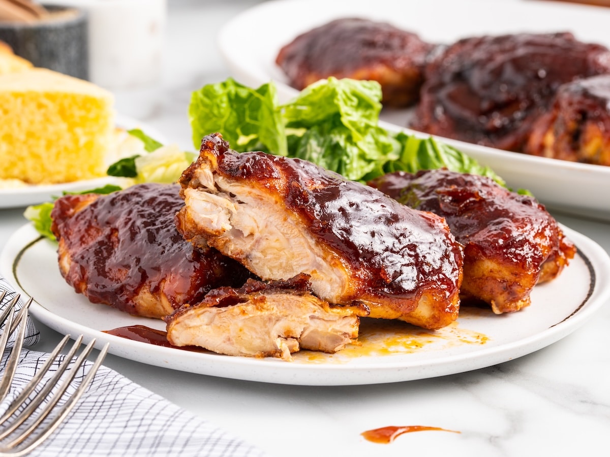 A plate of baked BBQ chicken thighs with one piece cut open to show juicy, fully cooked meat, served with lettuce and cornbread in the background.