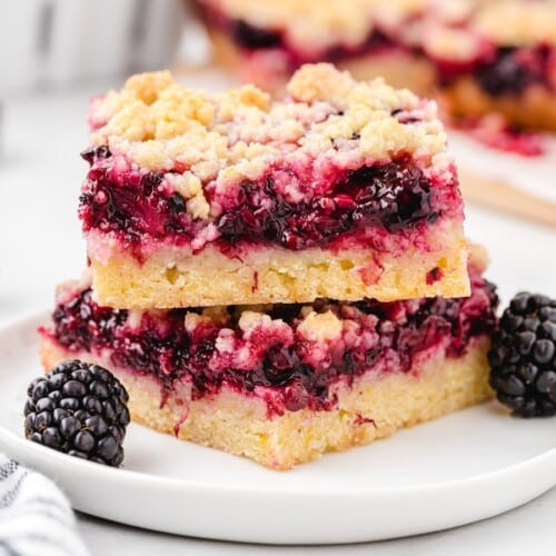 Close-up of blackberry bars stacked on a white plate, showing the buttery crust, dark berry filling, and golden crumble topping.