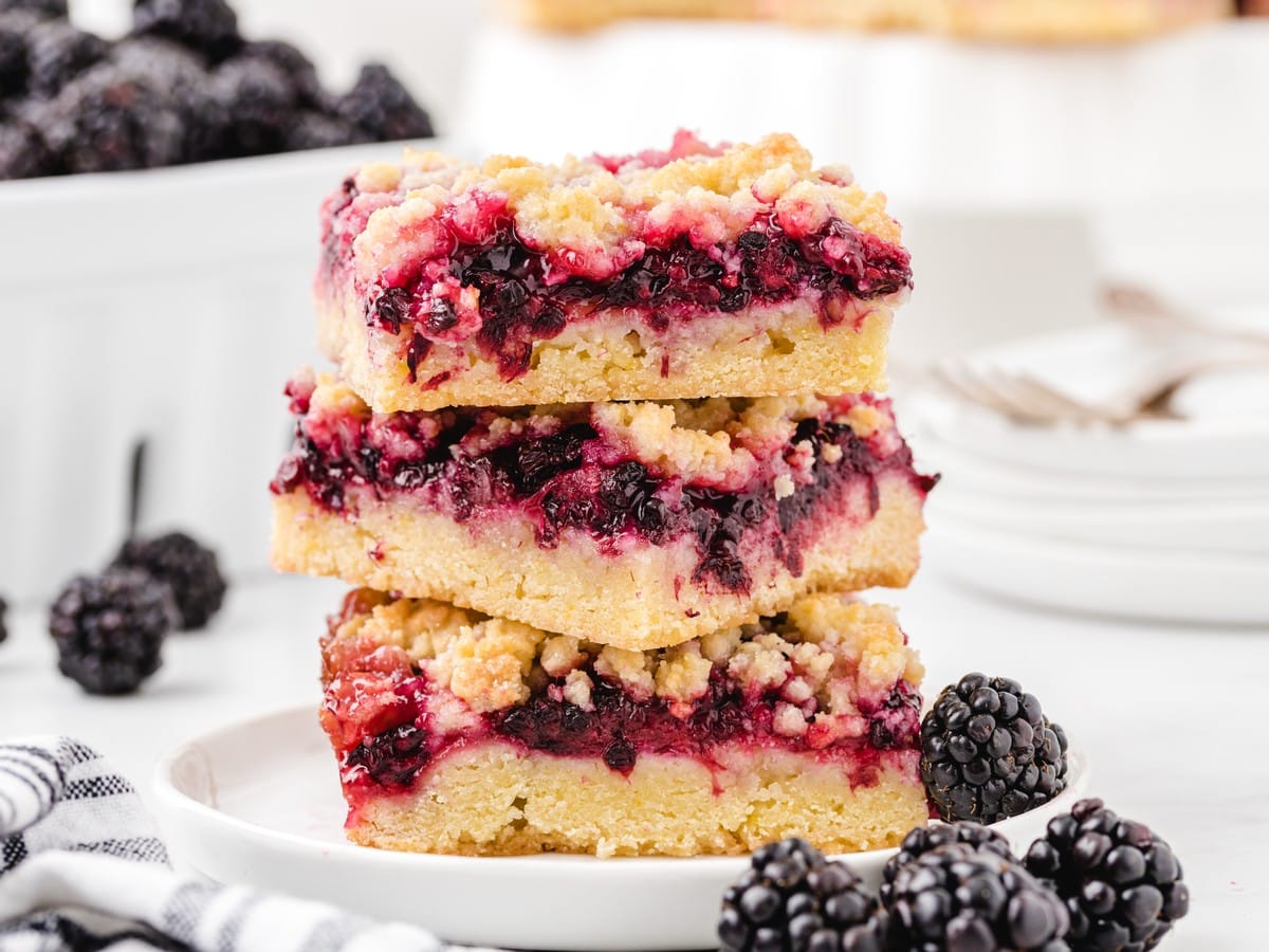 Three stacked blackberry bars with layers of buttery crust, glossy blackberry filling, and crumb topping, served with fresh blackberries.