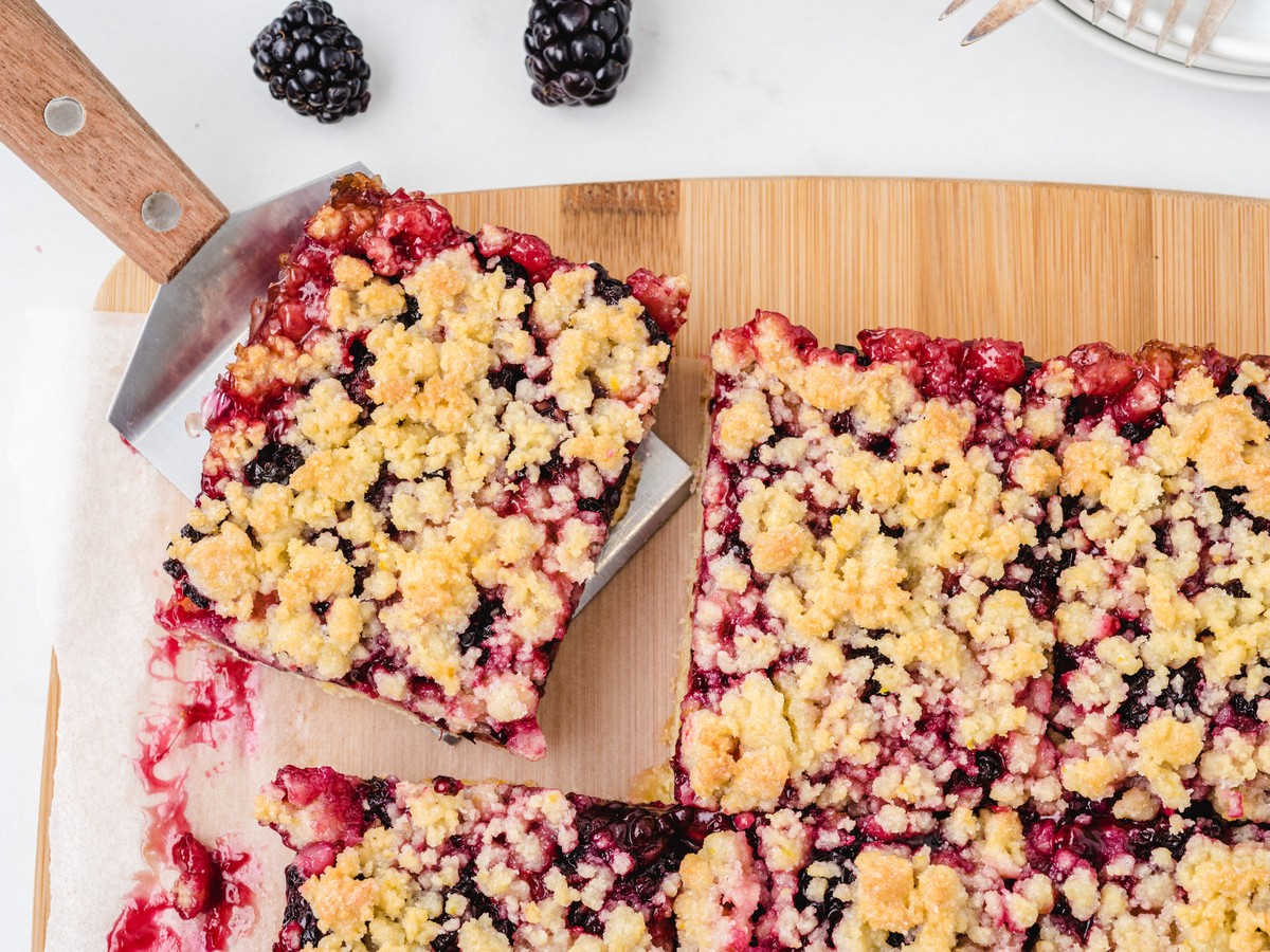 Overhead view of blackberry bars being lifted with a spatula, showing the golden crumb topping, blackberry filling, and parchment-lined pan.