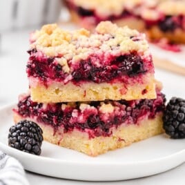 Stacked blackberry bars on a white plate with fresh blackberries, showing buttery shortbread crust, juicy blackberry filling, and golden crumb topping.