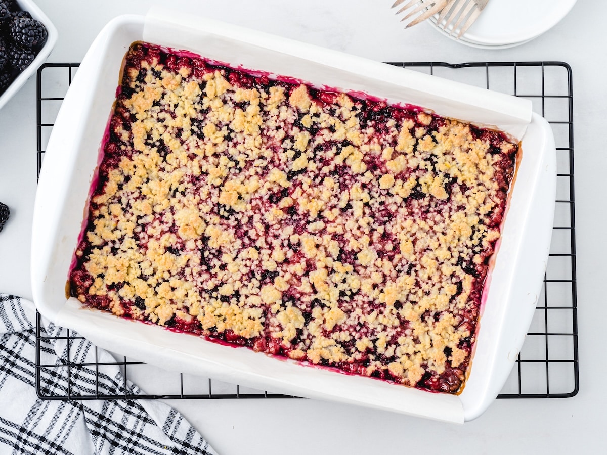 Baked blackberry pie bars with golden crumble topping in a white baking dish on a wire cooling rack