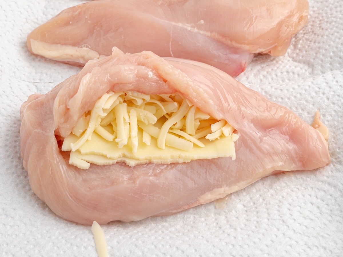 Close-up of a raw chicken breast stuffed with provolone and shredded mozzarella cheese on a paper towel-lined baking sheet.