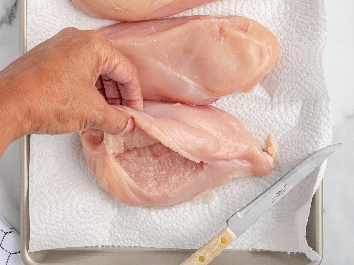 Hand holding open a pocket cut into a raw boneless skinless chicken breast on a paper towel-lined baking sheet beside a kitchen knife.