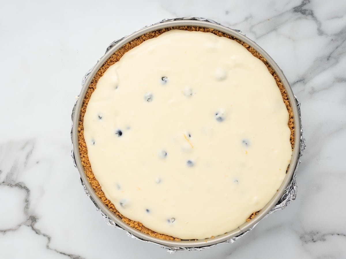 Lemon cheesecake batter with fresh blueberries folded in, spread evenly over the baked graham cracker crust in a foil-wrapped springform pan.