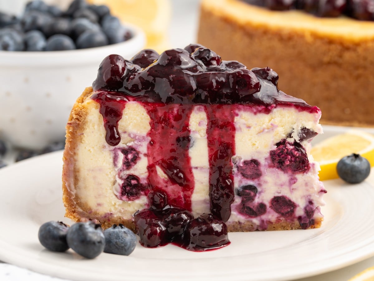 Single slice of lemon blueberry cheesecake topped with glossy blueberry sauce, with blueberry filling, graham cracker crust, fresh blueberries, and lemon slices.