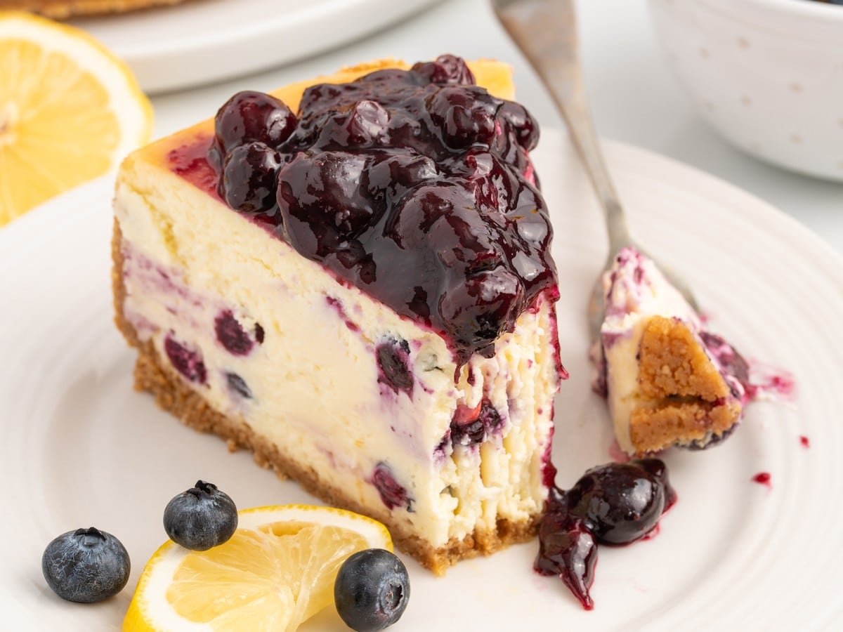 Slice of lemon blueberry cheesecake on a white plate with glossy blueberry topping, lemon slices, fresh blueberries, and a fork holding a bite.