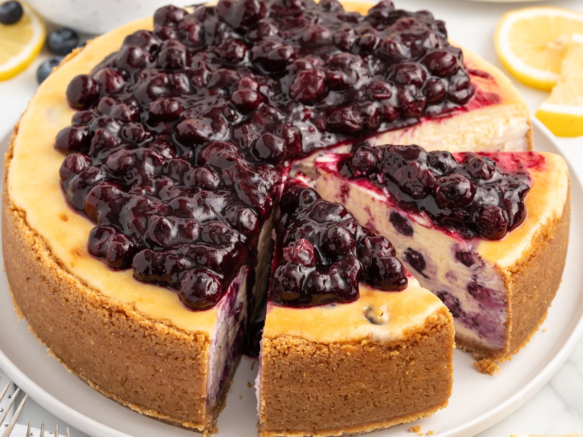 Whole lemon blueberry cheesecake with a graham cracker crust, glossy blueberry topping, lemon slices, and one slice cut from the cake.