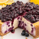 Whole lemon blueberry cheesecake with a slice removed showing blueberry-swirled filling, graham cracker crust, and glossy blueberry topping on a white plate with fresh blueberries.