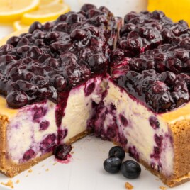 Whole lemon blueberry cheesecake with a slice removed showing blueberry-swirled filling, graham cracker crust, and glossy blueberry topping on a white plate with fresh blueberries.