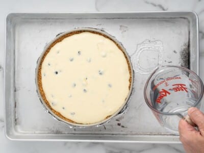 Pouring boiling water into a roasting pan around a foil-wrapped springform pan filled with lemon blueberry cheesecake batter.