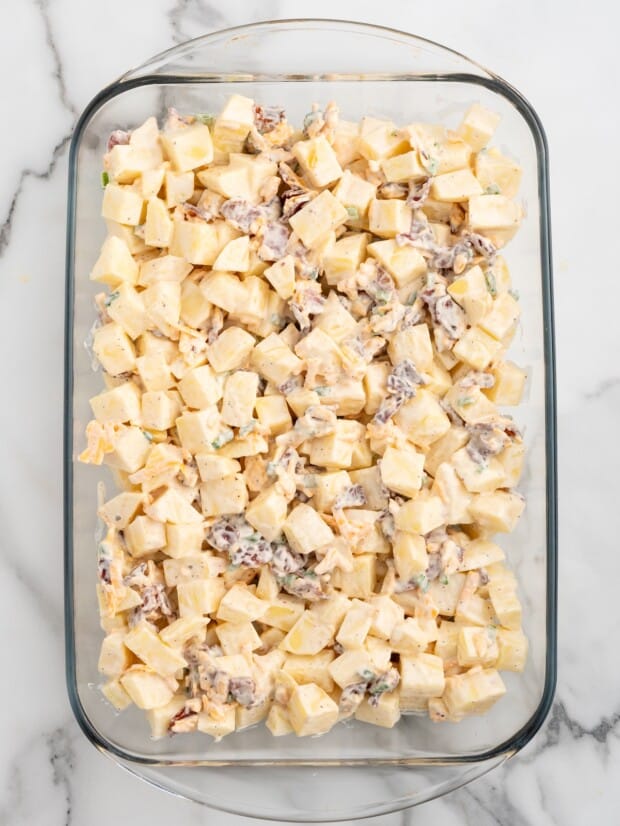 Raw diced Yukon gold potatoes coated in a seasoned mayonnaise mixture with bacon, cheddar cheese, and green onions in a glass 9x13 baking dish, ready to go into the oven.