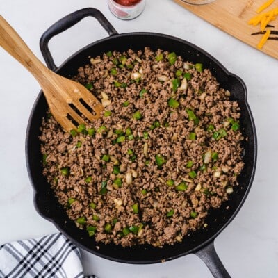 Ground beef, diced green pepper, and onion cooked together in a cast iron skillet, stirred with a wooden spoon.