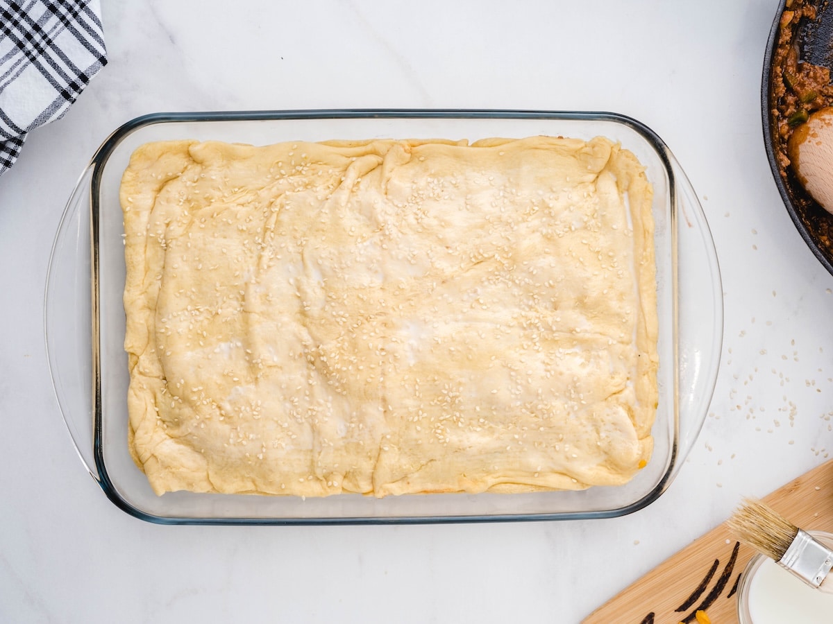 Second sheet of crescent roll dough laid over the filling in a glass baking dish, brushed with milk and sprinkled with sesame seeds, ready to bake.