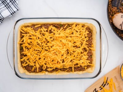 Sloppy joe filling spread over the pre-baked crescent roll crust in a glass baking dish, topped with a generous layer of shredded cheddar cheese.