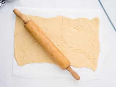 Crescent roll dough pressed and rolled flat on parchment paper with a wooden rolling pin, seam lines still visible across the surface.