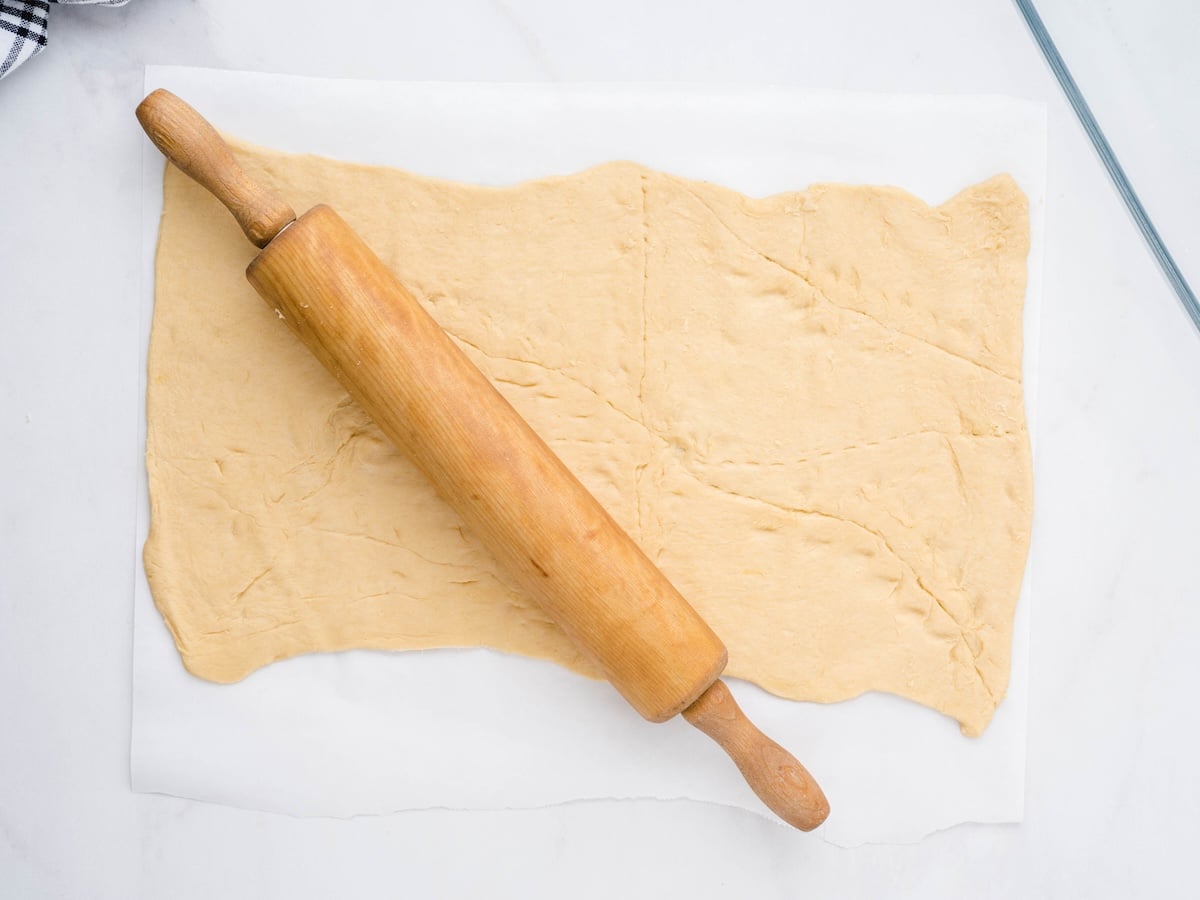 Crescent roll dough pressed and rolled flat on parchment paper with a wooden rolling pin, seam lines still visible across the surface.